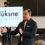 Četri cilvēki sēž pie galda sanāksmē. Priekšplānā pa kreisi vīrietis ar bārdu tumšā uzvalkā, centrā sirms vīrietis zilā uzvalkā ar saliktām rokām, un pa labi vīrietis tumšā uzvalkā ar gaišu kaklasaiti. Fonā sieviete baltā halātā. Uz galda ūdens krūze ar citronu un uzkodas.