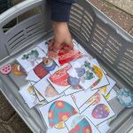 Bērna roka sniedzas pelēkā plastmasas grozā, kas ir pilns ar laminētām Lieldienu tematikas kartītēm. Kartītēs redzamas dekorētas Lieldienu olas un multfilmu truši ar skaitļiem, piemēram, "6" un "3", kas veido puzles gabaliņus.