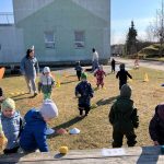 Bērna roka tumši zilā jakā sniedzas pelēkā grozā, kurā ir daudz Lieldienu puzles gabaliņu. Redzamas krāsainas olas ar ziediem, zvaigznēm un citiem rakstiem, kā arī zaķi, daži ar cipariem, piemēram, "3" un "6". Roka tūlīt paņems vienu gabaliņu.