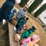 Bērna roka tumši zilā jakā sniedzas pelēkā plastmasas grozā, kas pilns ar dažādiem Lieldienu tēmas puzles gabaliņiem. Gabaliņos redzamas krāsainas, rotātas olas, karikatūru zaķi un vardes, un uz dažiem no tiem ir skaitļi, piemēram, "6" un "3".