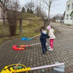 Bērna roka sniedzas pelēkā plastmasas grozā, kas ir pilns ar Lieldienu tēmas puzles gabaliem, tostarp krāsainām olām un trušiem. Roka tur puzles gabalu ar sarkanu olu un "X" rakstu, blakus gabalam ar ciparu "6".