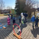 Bērna roka tumši zilā jakā sniedzas pelēkā grozā, kurā ir Lieldienu tēmas puzles gabaliņi ar krāsainām olām, zaķiem un cipariem, piemēram, 3 un 6.