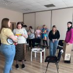 Sieviešu grupa gaišā telpā piedalās aktivitātē. Vairākas sievietes stāv, dažas tur priekšmetus: megafonu, papīrus un izrotātu kartona kasti. Viņas apspriežas un iesaistās.