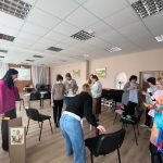 Sieviešu grupa piedalās nodarbībā iekštelpās. Priekšplānā sieviete tur skaļruni un sarkanu rotaļlietu, kamēr cita pēta dzeltenu papīra strēmeli. Pa labi no viņām sieviete rozā džemperī tur lielu kartona kasti, kas rotāta ar balta zaķim līdzīga tēla zīmējumu uz zirga. Fonā redzamas arī citas sievietes, kas iesaistītas aktivitātēs.