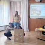 Sieviešu grupa piedalās iekštelpu darbnīcā. Priekšplānā sieviete dzeltenā kreklā tur rotaļlietu megafonu un runā ar citu sievieti, kura tur dzeltenu papīru. Labajā pusē sieviete tur lielu kartona kasti, uz kuras redzams zaķa zīmējums.