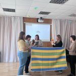 Sieviešu grupa piedalās iekštelpu aktivitātē. Priekšplānā sieviete dzeltenā kreklā tur megafonu, bet cita sieviete leoparda raksta svārkos pēta dzeltenu papīra strēmeli. Viņas t-krekla uzraksts ir "Feel your dreams". Pa labi sieviete tur lielu kartona kasti ar bruņinieka zīmējumu uz zirga. Citas sievietes fonā vēro.