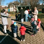 Sieviete tautas tērpā spēlē akordeonu bērnu un pieaugušo grupai saulainā dienā ārpus ēkas, uz bruģēta laukuma.