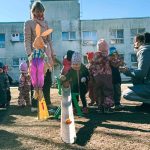 Gados veca sieviete tautas tērpā sēž ārā saulainā dienā un spēlē sarkanu akordeonu, kamēr bērnu grupa un pieaugušie stāv un klausās. Blakus viņai ir galds ar notīm un krāsainām bumbiņām.