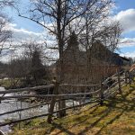 Saulaina pavasara dienas ainava ar akmeņainu upi kreisajā pusē un vienkāršu koka žogu. Labajā pusē redzama zālainā nogāze ar garām koku ēnām. Aiz kokiem bez lapām fonā redzama tumša, veca ēka zem zilu debesu ar mākoņiem.