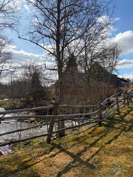 Saulaina pavasara dienas ainava ar akmeņainu upi kreisajā pusē un vienkāršu koka žogu. Labajā pusē redzama zālainā nogāze ar garām koku ēnām. Aiz kokiem bez lapām fonā redzama tumša, veca ēka zem zilu debesu ar mākoņiem.