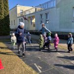 Bērna roka sniedzas pelēkā plastmasas grozā, kas pilns ar krāsainām, laminētām Lieldienu tēmas puzles detaļām. Detaļās redzamas dekorētas Lieldienu olas dažādos rakstos un krāsās, piemēram, tirkīza ar rozā ziediem un sarkana ar baltām zigzag līnijām, kā arī zaķi ar olu gabaliņiem, uz kuriem redzami cipari, piemēram, 3 un 6.