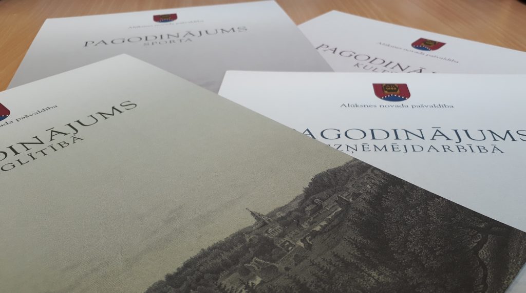 Vairāki baltas krāsas apbalvojumu dokumenti no Alūksnes novada pašvaldības, uz kuriem redzami uzraksti: "PAGODINĀJUMS SPORTĀ", "PAGODINĀJUMS KULTŪRĀ", "PAGODINĀJUMS IZGLĪTĪBĀ" un "PAGODINĀJUMS UZŅĒMĒJDARBĪBĀ". Priekšplānā esošajam dokumentam ir arī melnbalta pilsētas ilustrācija uz kalna.
