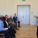 Telpā ar koka grīdu sieviete rūtainā žaketē runā un žestikulē ar rokām, vēršoties pie sēdošas auditorijas. Auditorijā ir gan jaunas sievietes, gan vīrieši militārajās formās. Aizmugurē redzamas baltas durvis, zaļa avārijas izejas zīme un klavieres ar uzrakstu "KAWAI".