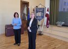 Divas sievietes stāv iekštelpās; viena runā, bet otra tur rokās nelielu priekšmetu. Fonā redzams zils karogs ar skolas ēkas attēlu un tekstu "ERNSTA GLIKA ALŪKSNES VALSTS ĢIMNĀZIJA", Latvijas un Eiropas Savienības karogi, kā arī liels ekrāns ar ēkas un zaļumu attēlu.