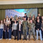 Kopīga fotogrāfija ar aptuveni trīsdesmit skolēniem, skolotājiem, vīrieti uzvalkā un divām militārpersonām, kas stāv iekštelpās pie skatuves. Fonā liels ekrāns ar skolas ēkas attēlu. Kreisajā pusē redzams baneris ar uzrakstu "ERNSTA GLIKA ALŪKSNES VALSTS ĢIMNĀZIJA", bet labajā pusē – Latvijas karogs. Priekšplānā viens no skolēniem valkā džemperi ar uzrakstu.