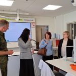 Četri cilvēki sarunājas ķīmijas laboratorijā. Vīrietis militārajā formā ar uzšuvi, sieviete pelēkā žaketē, sieviete gaiši zilā džemperī un sieviete melnā žaketē stāv pie galdiem ar laboratorijas aprīkojumu. Fonā redzama tāfele ar ķīmiskām formulām un elementiem "K, Na, Li, Ba, Sr, Ca, Mg, Ar", "Pb, H, Cu, Hg, Ag, Pt, Au", kā arī plakāti.