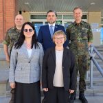 Pieci cilvēki – divas sievietes priekšplānā un trīs vīrieši aizmugurē, divi militārajās uniformās un viens uzvalkā – pozē grupas fotogrāfijai uz āra kāpnēm pie ēkas.