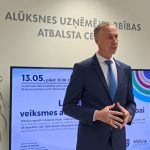 Vīrietis tumšā uzvalkā un zilā kaklasaitē runā un žestikulē ar rokām pasākumā. Aiz viņa ir siena ar uzrakstu "ALŪKSNES UZŅĒMĒJDARBĪBAS ATBALSTA CENTRS" un liels ekrāns, kurā redzama informācija par semināru: "13.05. plkst. 11.00-17" "Uzņēmējdarbības atbalsta centra telpās" "Alūksnes kultūras centrs, Brūža iela 7". Uz ekrāna tiek runāts par atbalstu, ko var saņemt uzņēmēji Alūksnes novadā. Redzams arī Alūksnes logo.