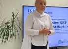 Sieviete baltā kreklā un tumšās biksēs stāv un runā seminārā. Fonā uz sienas redzams uzraksts "ALŪKSNES UZNĒMĒJDA ATBALSTA CENTRS". Uz ekrāna aiz viņas ir redzami vārdi "informatīvs seminārs" un "Latgales SEZ", kā arī informācija par nodokļu atvieglojumiem uzņēmējiem speciālās ekonomiskās zonas ietvaros un "LATGA" logotips.