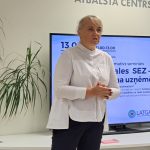 Sieviete baltā kreklā un tumšās biksēs stāv un runā seminārā. Fonā uz sienas redzams uzraksts "ALŪKSNES UZNĒMĒJDARBĪBAS ATBALSTA CENTRS". Uz ekrāna aiz viņas ir redzami vārdi "informatīvs seminārs" un "Latgales SEZ", kā arī informācija par nodokļu atvieglojumiem uzņēmējiem speciālās ekonomiskās zonas ietvaros.