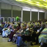 Cilvēki sēž modernā auditorijā. Daudzi tur ziedu pušķus, un virs krēsliem peld zaļš balons. Zāli izgaismo skatuves prožektori un dekoratīvais griestu apgaismojums, radot svinīgu atmosfēru.