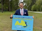 Smaidošs vīrietis uzvalkā un kaklasaitē tur zilu plakātu ar tekstu "BEZPROCENTU AIZDEVUMS biznesa izaugsmei Latvijas austrumu pierobežā". Plakātā ir Latvijas karte ar iezīmētiem novadiem (Alūksnes, Balvu, Ludzas, Krāslavas, Augšdaugavas) un papildu informāciju: "0 % likme bez EURIBOR līdz 01.01.2029. Piesakoties jaunām investīciju vai apgrozāmo līdzekļu aizdevumam līdz 30.06.2027.". Vīrietis ar otru roku rāda īkšķi uz augšu, fonā redzami koki un zāle.