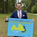 Smaidošs vīrietis uzvalkā un kaklasaitē tur zilu plakātu ar tekstu "BEZPROCENTU AIZDEVUMS biznesa izaugsmei Latvijas austrumu pierobežā". Plakātā ir Latvijas karte ar iezīmētiem novadiem (Alūksnes, Balvu, Ludzas, Krāslavas, Augšdaugavas) un papildu informāciju: "0 % likme bez EURIBOR līdz 01.01.2029. Piesakoties jaunām investīciju vai apgrozāmo līdzekļu aizdevumam līdz 30.06.2027.". Vīrietis ar otru roku rāda īkšķi uz augšu, fonā redzami koki un zāle.