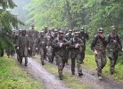 Grupa militārpersonu kamuflāžas tērpos lietū soļo pa slapju dubļainu taku cauri zaļam mežam. Divas sievietes skatās savās rokas ierīcēs.