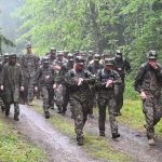 Grupa militārpersonu kamuflāžas tērpos lietū soļo pa slapju dubļainu taku cauri zaļam mežam. Divas sievietes skatās savās rokas ierīcēs.