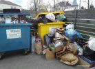 Divi pārpildīti šķirošanas konteineri, viens zils un viens dzeltens, ar uzrakstu "PLASTMASAS, PAPĪRA, METĀLA IEPAKOJUMI". Ap konteineriem uz zemes sakrauti daudz atkritumu, tostarp kartona kastes un plastmasas maisi.