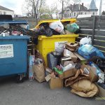 Divi pārpildīti šķirošanas konteineri, viens zils un viens dzeltens, ar uzrakstu "PLASTMASAS, PAPĪRA, METĀLA IEPAKOJUMI". Ap konteineriem uz zemes sakrauti daudz atkritumu, tostarp kartona kastes un plastmasas maisi.