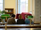 Trīs cilvēki, divas sievietes un vīrietis, sēž pie galda, skatoties klēpjdatorā bibliotēkas lasītavā. Fonā redzams grāmatu plaukts un logi, priekšplānā – zaļas galda lampas.