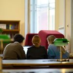 Trīs cilvēki, divas sievietes un vīrietis, sēž pie galda, skatoties klēpjdatorā bibliotēkas lasītavā. Fonā redzams grāmatu plaukts un logi, priekšplānā – zaļas galda lampas.