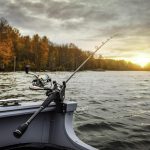 Makšķere ar spoli piestiprināta laivas sānā, metoties viļņainā ezerā saulrietā. Krastu rotā rudens koki zem daļēji mākoņainām debesīm ar zelta saules spīdumu.