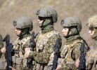Vairāki karavīri, tostarp sievietes, kamuflāžas uniformās, taktisko vestēs un ķiverēs stāv rindā, turot šautenes.