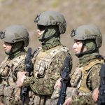Vairāki karavīri, tostarp sievietes, kamuflāžas uniformās, taktisko vestēs un ķiverēs stāv rindā, turot šautenes.