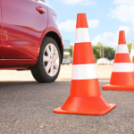 Sarkana automašīna stāv blakus oranžu un baltu satiksmes konusu rindai uz asfalta, saulainā dienā, kas norāda uz braukšanas nodarbību vai eksāmenu.