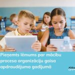 Divi bērni sēž klasē un koncentrēti skatās savos planšetdatoros. Attēlā redzamais teksts: Pieņemts lēmums par mācību procesa organizāciju gaisa apdraudējuma gadījumā. Alūksne Oinkaua.