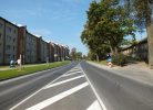 Pilsētas iela saulainā dienā ar daudzdzīvokļu namiem kreisajā pusē un vecākām koka ēkām labajā pusē. Pa ietvi iet cilvēks, stumjot bērnu ratiņus. Redzamas baltas ceļa marķējuma līnijas un ceļa zīmes, kas norāda līkumu un apstāšanās aizliegumu.