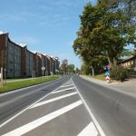Pilsētas iela saulainā dienā ar daudzdzīvokļu namiem kreisajā pusē un vecākām koka ēkām labajā pusē. Pa ietvi iet cilvēks, stumjot bērnu ratiņus. Redzamas baltas ceļa marķējuma līnijas un ceļa zīmes, kas norāda līkumu un apstāšanās aizliegumu.