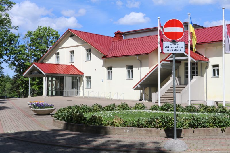 Gaiši dzeltena ēka ar sarkanu jumtu un vairākām ieejām. Priekšplānā bruģēts celiņš un apstādījumi. Pie ēkas redzama zīme "Iebraukt aizliegts" ar papildtekstu "Izņemot ar Alūksnes kultūras centra atļauju" un mastos plīvo Latvijas un Ukrainas karogi.