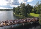 Aerofotogrāfija ar lielu cilvēku grupu, kas pulcējas uz koka tilta virs ūdens, ko ieskauj zaļi koki un zālāji zem daļēji mākoņainām debesīm.
