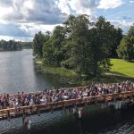 Aerofotogrāfija ar lielu cilvēku grupu, kas pulcējas uz koka tilta virs ūdens, ko ieskauj zaļi koki un zālāji zem daļēji mākoņainām debesīm.