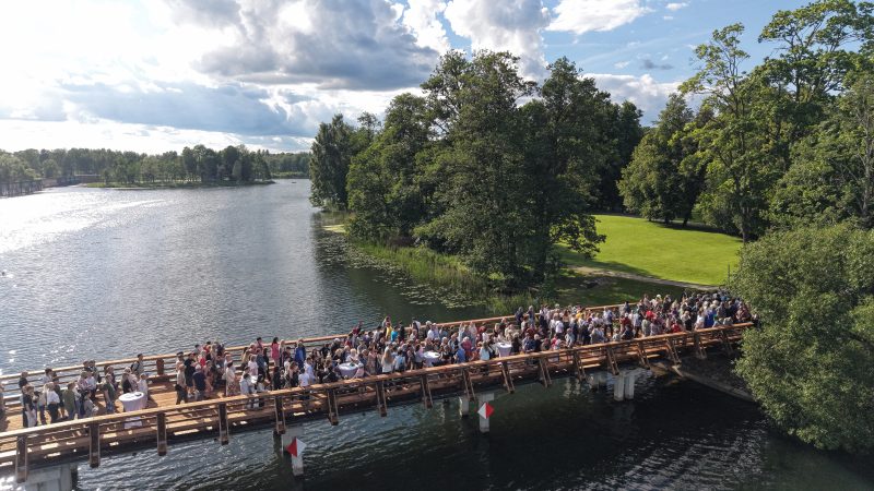 Aerofotogrāfija ar lielu cilvēku grupu, kas pulcējas uz koka tilta virs ūdens, ko ieskauj zaļi koki un zālāji zem daļēji mākoņainām debesīm.