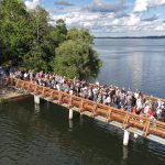 Panorāmas skats uz garu koka tiltu, kas pārpildīts ar cilvēkiem, pār ezeru, savienojoties ar zaļu, kokiem apaugušu krastu. Ezerā redzamas laivas, viena no tām veido ūdens taku zem daļēji mākoņainām debesīm.