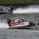 Divas ātrgaitas laivas sacīkstēs uz ūdens. Priekšplānā melna, oranža un balta laiva ar numuru "51" un uzrakstiem "MERCURY", "F1POWERBOAT", "MEMELES" un "22448254", veidojot lielas šļakatas. Aizmugurē pelēka laiva ar numuru "22" un "MERCURY" uz dzinēja.