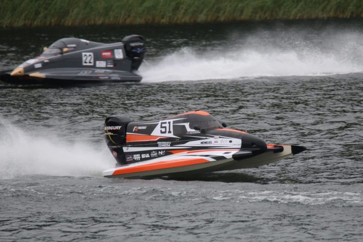 Divas ātrgaitas laivas sacīkstēs uz ūdens. Priekšplānā melna, oranža un balta laiva ar numuru "51" un uzrakstiem "MERCURY", "F1POWERBOAT", "MEMELES" un "22448254", veidojot lielas šļakatas. Aizmugurē pelēka laiva ar numuru "22" un "MERCURY" uz dzinēja.