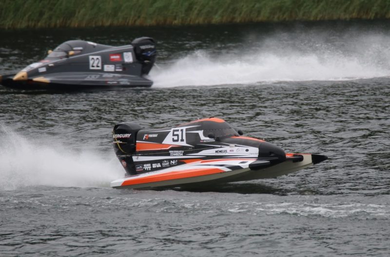 Divas ātrgaitas laivas sacīkstēs uz ūdens. Priekšplānā melna, oranža un balta laiva ar numuru "51" un uzrakstiem "MERCURY", "F1POWERBOAT", "MEMELES" un "22448254", veidojot lielas šļakatas. Aizmugurē pelēka laiva ar numuru "22" un "MERCURY" uz dzinēja.