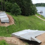 Divas jaunas betona konstrukcijas smilšainā, zālainā nogāzē, blakus ezeram. Augšējā ir gara, slīpa betona rampa, bet apakšējā — platāka, daudzstūra formas betona platforma. Fonā redzams ezers ar koka piestātni un lapeni, kā arī zaļš krasts ar celiņu un kokiem.