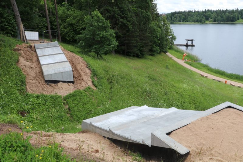 Divas jaunas betona konstrukcijas smilšainā, zālainā nogāzē, blakus ezeram. Augšējā ir gara, slīpa betona rampa, bet apakšējā — platāka, daudzstūra formas betona platforma. Fonā redzams ezers ar koka piestātni un lapeni, kā arī zaļš krasts ar celiņu un kokiem.